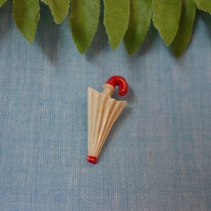 May include: A miniature umbrella figurine with a cream-colored canopy and a red handle and tip. The umbrella is set against a light blue textured background, with green leaves at the top.