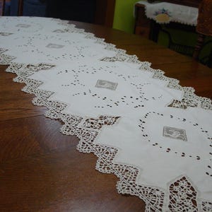 May include: A long white table runner featuring intricate cutwork embroidery and a wide, decorative lace border. The runner is composed of several square panels, each centered with a small, square-shaped beige embroidered motif of a bird.