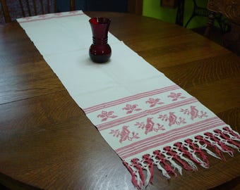May include: A long, rectangular off-white table runner featuring red embroidered bird and floral motifs at both ends, finished with a fringe of red and white threads. A small, dark red glass vase sits in the center of the runner.