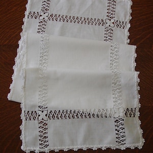 May include: White linen placemats with intricate lace detailing. The placemats feature a rectangular shape with a cross-shaped lace design in the center and along the edges. The lace is delicate and adds a touch of elegance to the simple design.