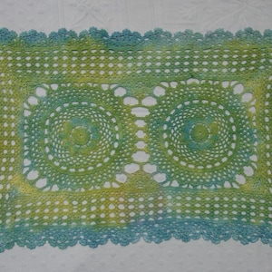 May include: Handmade crochet doily in shades of green, yellow, and blue. The rectangular doily features a central design of two circular patterns, surrounded by a lacy border. The edges are finished with a scalloped trim.
