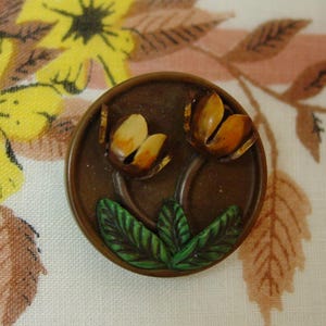 May include: A round, brown button with a floral design. Two stylized flowers with yellow and orange petals are depicted, along with green leaves. The button is likely made of plastic or resin and is suitable for sewing or crafts.