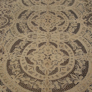 May include: A beige lace tablecloth featuring a repeating pattern of large circular medallions connected by intricate floral and vine motifs. The design is composed of openwork mesh and solid thread elements, creating a detailed, symmetrical textile.