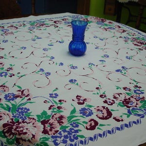 May include: A vintage white tablecloth with a floral pattern in shades of pink, purple, and blue. The tablecloth has a scalloped border and a blue glass vase in the center. The floral design includes large blooms and delicate swirls.
