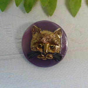 May include: A circular button featuring a purple base with a raised, metallic gold-toned cat face relief in the center. The cat has detailed fur texture, pointed ears, and prominent black whiskers extending from the nose area across the purple surface.