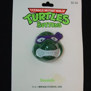 May include: Teenage Mutant Ninja Turtles button featuring Donatello, the purple masked turtle with a green shell and white eyes. The button is on a white card with the Teenage Mutant Ninja Turtles logo and the text "$2.50".
