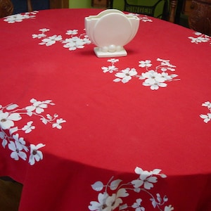 May include: A red oval tablecloth with a white floral pattern and a white ceramic vase. The tablecloth is made of a solid red fabric with white flowers and leaves scattered across the surface.