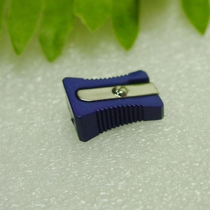 May include: A small, dark blue pencil sharpener with a silver blade. The sharpener has a rectangular shape with ridged sides for grip. The background is a light, textured surface with green leaves in the upper left corner.