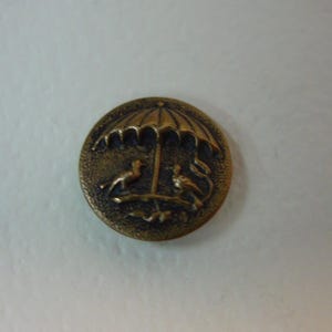 May include: A round, antique brass button with an umbrella design. Two birds are depicted under the umbrella. The button has a detailed, embossed design and a slightly tarnished finish.
