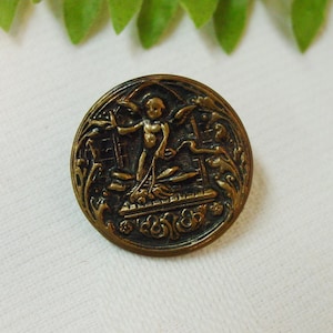 May include: A vintage brass button with an ornate design featuring a cherub figure with wings, surrounded by floral motifs. The button has a dark patina and a raised design.