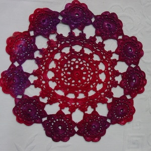 May include: A crocheted doily with a red and purple ombre design. The doily is circular and has a delicate lace pattern.