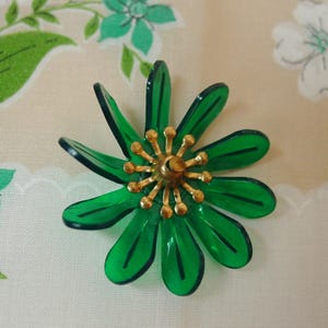 May include: A vintage flower brooch with a gold-tone center and translucent green petals. The brooch has a detailed design, with each petal and the center's details clearly visible. The brooch is a floral accessory.