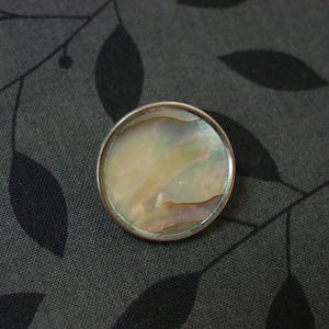 May include: A round, iridescent mother-of-pearl cabochon set in a silver-colored metal bezel. The cabochon has a pearlescent sheen with hues of cream, gold, and pink. The jewelry piece is set against a dark gray fabric with a leaf pattern.