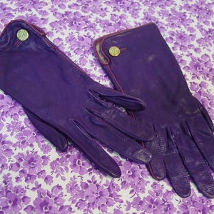 May include: A pair of purple leather gloves featuring a smooth finish, a rounded cuff design, and a single gold-toned circular button detail on the wrist of each glove. The gloves have visible stitching along the edges and fingers.