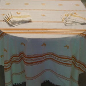 May include: A white tablecloth with horizontal orange stripes and small embroidered yellow bird motifs, accompanied by two stacks of matching napkins. The fabric features a fringed edge along the bottom and a woven texture throughout the set.