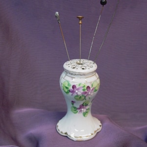 May include: A white ceramic pin cushion holder with a flared base and a perforated top, decorated with hand-painted purple violet flowers and green leaves. Four long metal hat pins with varied decorative heads are inserted into the top holes.
