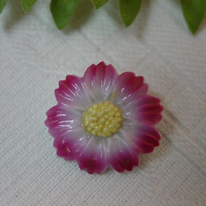 May include: A close-up of a flower-shaped brooch. The brooch has a white center with a yellow textured center. The petals are a gradient of pink to white. The brooch is on a white textured surface.