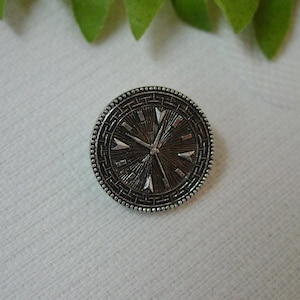 May include: A round, silver-toned button with a clock face design. The button features a detailed pattern around the edge and a textured center with radiating lines and arrow-shaped markers. The button is approximately 1 inch in diameter.