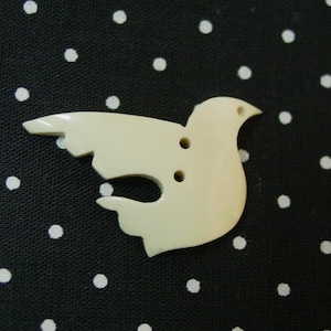 May include: A white, ceramic button shaped like a dove with two holes for sewing. The button is on a black background with white polka dots.