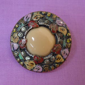 May include: A round brooch featuring a smooth, beige, dome-shaped center surrounded by a dark metal frame. The frame is decorated with painted leaves in shades of red, yellow, and lavender, interspersed with small, clear, sparkling rhinestones.