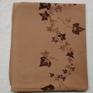 May include: Folded tan fabric with a repeating pattern of dark brown and light brown ivy leaves and vines. The leaves are stylized and arranged in a flowing, organic design. The fabric appears to be a medium weight material.