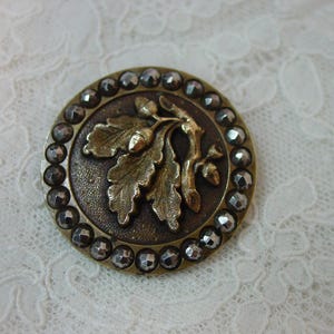 May include: A round, antique button with a bronze-colored base and a detailed oak leaf and acorn design in gold. The button is encircled by a row of faceted, silver-colored stones, adding a touch of sparkle.
