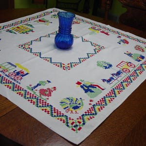 May include: A white square tablecloth with colorful embroidered designs and a blue glass vase. The embroidery features figures, vehicles, and geometric patterns in red, green, blue, and yellow. The vase is a deep blue color.