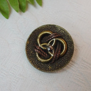 May include: A round, dark bronze-toned button featuring a textured, speckled surface. The center displays an intricate, raised design of intertwined metallic loops in a brass finish, accented with a small, clear, faceted crystal at the very middle.
