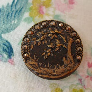 May include: A round, dark brown button featuring a raised, embossed design of a tree with leaves and grass. The outer edge of the button is decorated with a series of small, metallic, faceted studs arranged in a circular pattern around the center.