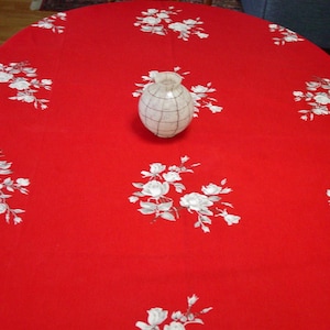 May include: Red oval tablecloth with a white floral pattern. A white vase with a textured surface sits in the center of the tablecloth.
