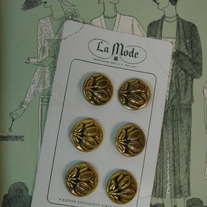 May include: A card of six vintage gold-tone buttons with a floral design. The card is labeled "La Mode" and includes the text "Button Specialists Since 1877. Superior Quality. Made in U.S.A." The background features illustrations of women in vintage clothing.