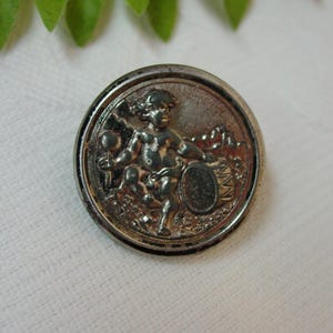 May include: A silver-toned button with a raised design of a cherub playing a drum. The button has a smooth, shiny surface.