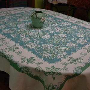 May include: A rectangular tablecloth featuring a white base with a large central panel of teal and white floral patterns, surrounded by a scalloped border of green and white leaves and flowers, with a light green ceramic vase placed on top.