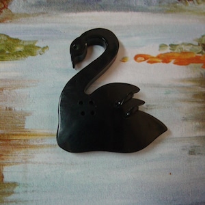 May include: A black swan-shaped decorative object. The swan has a curved neck and detailed wings. The object is made of a glossy material and has a smooth finish. The swan is set against a background with a water and landscape design.