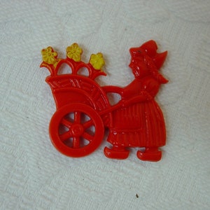 May include: A red plastic figurine of a girl pushing a red wagon with yellow flowers. The girl is wearing a red dress and a red hat.