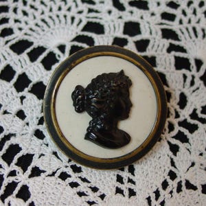 May include: A round, vintage brooch with a black silhouette of a woman's profile on a white background. The brooch has a black and gold-toned metal frame. The background is a white, lacy fabric.