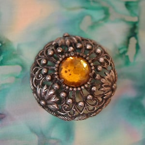 May include: A round silver-toned metal brooch featuring an intricate filigree pattern with small raised spheres and leaf motifs. A single, large, faceted amber-colored glass stone is set securely in the center of the circular piece.
