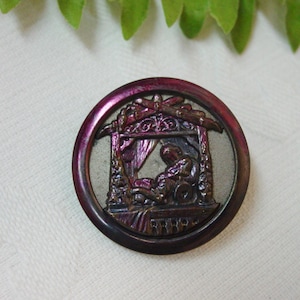 May include: A round, antique button with a detailed relief scene. The button has a dark, iridescent outer ring and a silver-toned center. The relief depicts a figure in a draped setting, possibly a religious or historical scene.