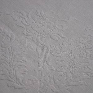 May include: A white textile featuring a tone-on-tone damask weave. The fabric displays an intricate, raised pattern of a central floral bouquet contained within a vase, flanked by symmetrical, elaborate scrolling acanthus leaf motifs.
