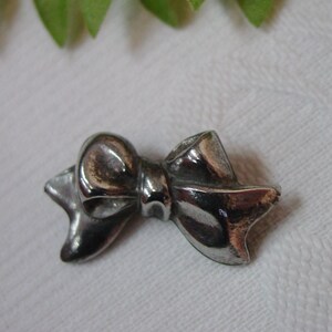 May include: A small, silver-colored bow-shaped charm. The charm has a polished, reflective surface and is set against a white background. The bow's design includes a central knot and looped ends.