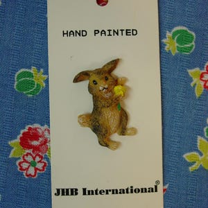 May include: A brown rabbit button with a white tail, holding a small yellow flower. The button is attached to a white rectangular card with the text HAND PAINTED at the top and JHB International at the bottom. The rabbit has dark brown ears.