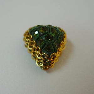 May include: A gold-toned, triangular-shaped brooch with a chain-link border. The brooch is encrusted with green, faceted gemstones. The gemstones are arranged in a geometric pattern, creating a textured surface. The brooch is set against a plain white background.