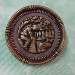 May include: A gold-toned metal button with a raised design of a person sitting on a wall with a bird in the background. The button has a decorative border.