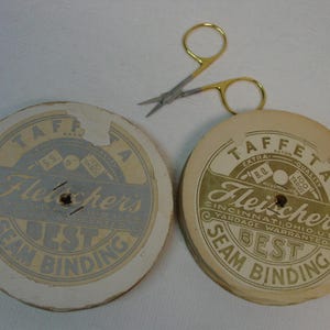 May include: Two round cardboard spools of Fleisher's Best Seam Binding, one with a blue label and one with a tan label, each marked 100 yards. A pair of small sewing scissors with gold-colored handles and silver blades rests above the spools.