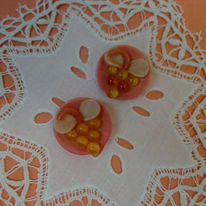 May include: Two circular buttons featuring a pink base with a cluster of small, translucent amber-colored beads and two smooth, pale beige petal-like shapes attached to the top, creating a decorative fruit-inspired design on each piece.