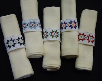 May include: Five white linen napkins with embroidered star-shaped napkin rings. The rings are in different colors: black, green, blue, red, and white.