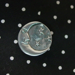 May include: A silver-colored celestial-themed button featuring a crescent moon with a face and three stars. The button is round and sits on a black fabric with white polka dots. The button is a decorative accessory.
