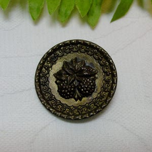 May include: A round, brass button with a raised design of two berries and a leaf. The button has a decorative border.