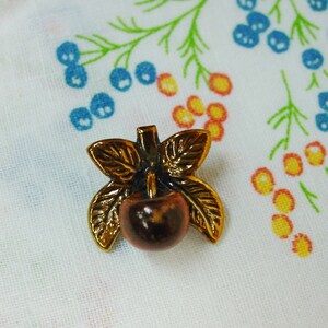 May include: A gold-toned metal button with a brown cherry and two gold leaves. The cherry is in the center of the button and the leaves are on either side of the cherry.