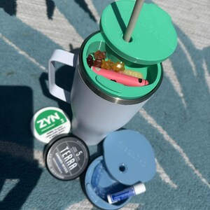 Puede incluir: Un vaso aislado blanco con una tapa verde y una pajita gris. La tapa está parcialmente abierta, revelando caramelos de goma y un artículo rosa. También son visibles dos recipientes pequeños y un recipiente azul con un bálsamo labial.
